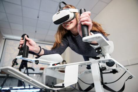 Foto von einer Person auf dem Icaros-Gerät. Sie trägt eine VR-Brille und hält sich an den Griffen fest.