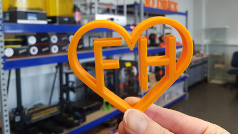 Eine Hand hält ein orangefarbenes Herz aus dem 3D-Drucker, das die Buchstaben F und H umschließt. Im Hintergrund stehen Regale mit Geräten.