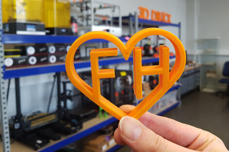 One hand holds an orange heart from the 3D printer, which encloses the letters F and H. In the background are shelves with appliances.