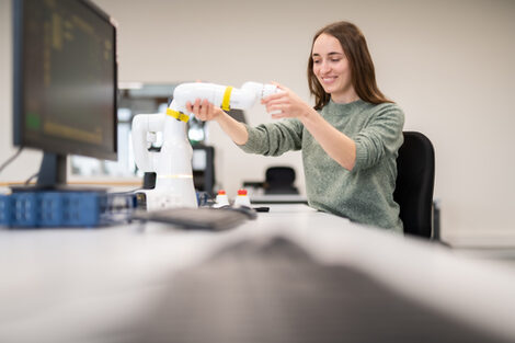 Foto einer Studierenden, die an einem Computertisch sitzt und mit der Hand den Arm eines Schrenkarmroboters hält.