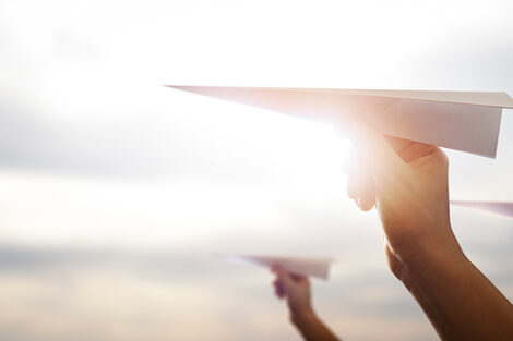 Photo of three hands each holding a paper airplane in front of a sunny sky.