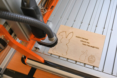 Auf einem Lasercutter liegt eine dünne Holzplatte mit den Konturen von zwei Hasen, einem Korb mit Eiern sowie dem Satz: Schöne Feiertage wünscht das Team FabLab@FHDO.