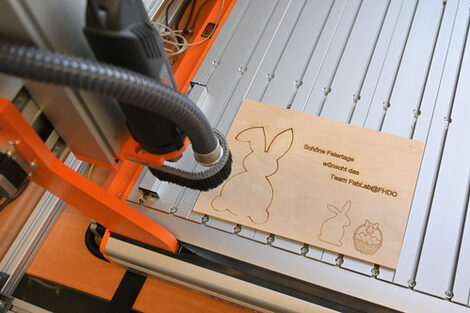 On a laser cutter lies a thin wooden plate with the outlines of two rabbits, a basket with eggs and the sentence: Happy holidays from the FabLab@FHDO team.