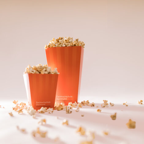 Two popcorn bags, one large and one small, are dramatically illuminated from behind. Popcorn lies around them. The bags stand on a white background.