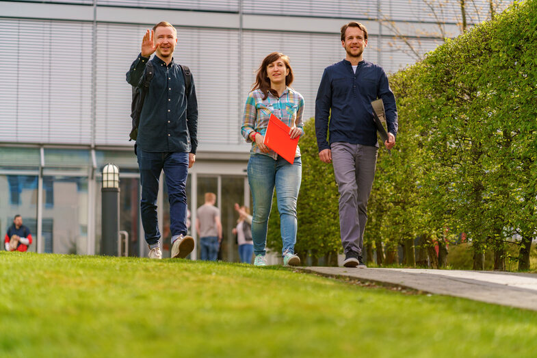 Foto von einer Studentin und zwei Studenten, die nebeneinander über den Campus gehen. Der eine Student winkt jemandem zu.__Three students walk side by side on a path, the student on the left raises his hand in greeting.