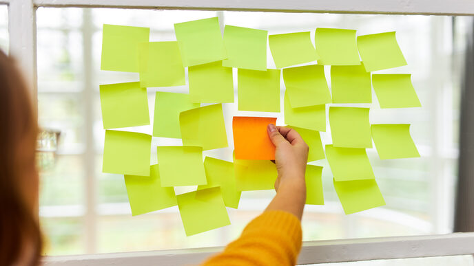 Foto von vielen gelben Post-its, die neben- und untereinander an einer Scheibe kleben. In der Mitte klebt ein einziges orangefarbenes Post-it. Eine Frau steht davor und greift nach dem orangenen Post-it.