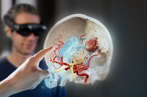 Ein junger Mann im Hintergrund sieht sich durch eine Virtual Reality Brille den Querschnitt von Hirnschichten mit verschiedenen anatomischen Strukturen an. Am linken Bildrand erscheint eine Hand im Vordergrund, die mit dem Zeigefinger auf das Hologramm zeigt.