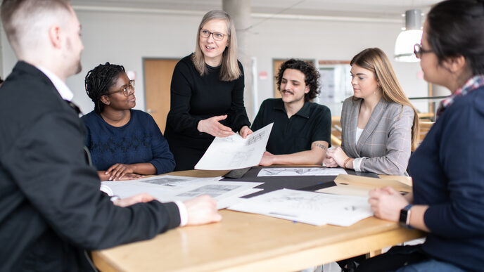 Photo of an employee of the Faculty of Architecture. She is at a table with drawings on it, surrounded by students. She is holding a drawing in her hand and pointing to it with her other hand, smiling as she looks at a student sitting to the left of the picture. __ <br>An employee of the architecture department stands at a table with drawings on it, surrounded by students. She holds a drawing in her hand, which she points to with the other hand, and she smiles and looks at a student who is sitting in the left corner of the picture.