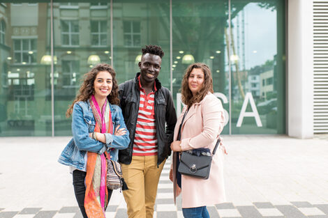 Foto von zwei Studentinnen und einem Studenten, die nebeneinander vor dem Mensagebäude stehen. Sie lachen in die Kamera.__Two female and one male students stand side by side in front of the canteen building. They laugh at the camera.