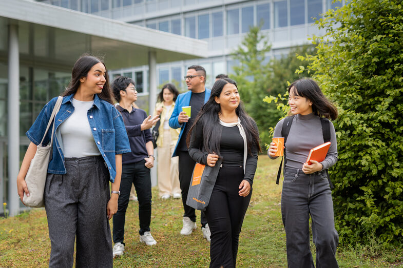 Group of international students walking together across a meadow on the Emil-Figge-Straße campus with bags and drinks cups in front of university buildings.
