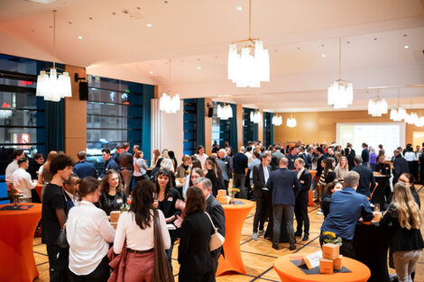 One room is festively lit with ceiling lamps, several people stand in groups at tables and chat.