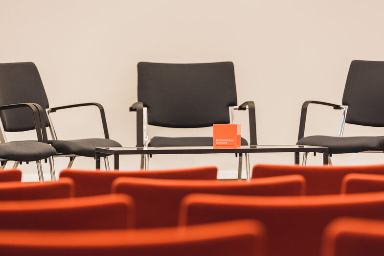 Photo of an empty event room, view over empty rows of chairs to the podium, where there are more chairs and a table with FH cube. __ Empty event room, view over empty rows of chairs to the podium, where there are more chairs and a table with FH cube.
