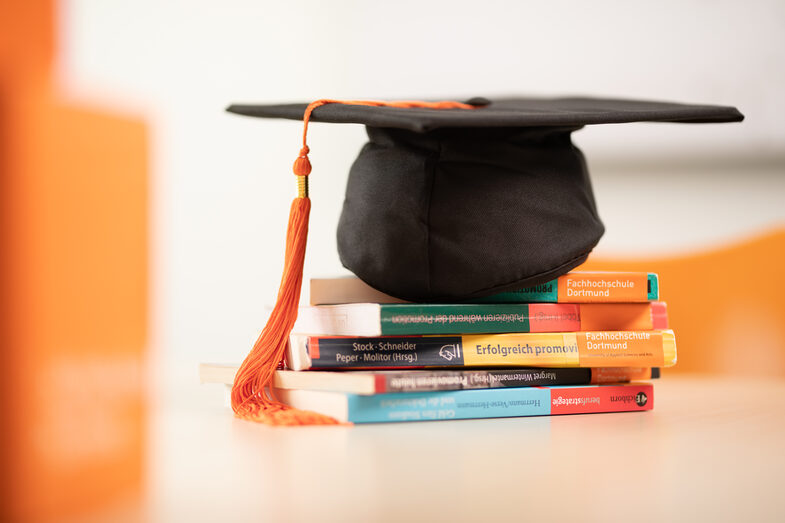 Foto eines Doktorhuts, der auf einem Bücherstapel bestehend aus Ratgebern zur Promotion liegt.