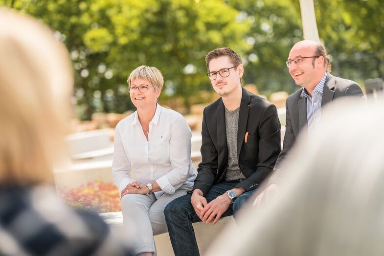 Foto von drei Personen, die draußen auf dem Campus sitzen. Ein Promovierender in der Mitte, daneben zwei Kollegen, die ihn bei der Promotion betreuen.