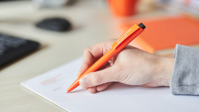 Foto einer Hand, die mit einem Kugelschreiber auf einen Notizblock schreibt. Der Kugelschreiber ist orange und trägt den Schriftzug der Fachhochschule in weiß.