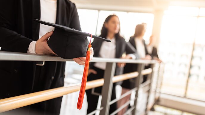 Photo of four doctoral students - blurred - standing at a railing in the building. The focus is on a doctor's hat, which one of them is holding in his hand.