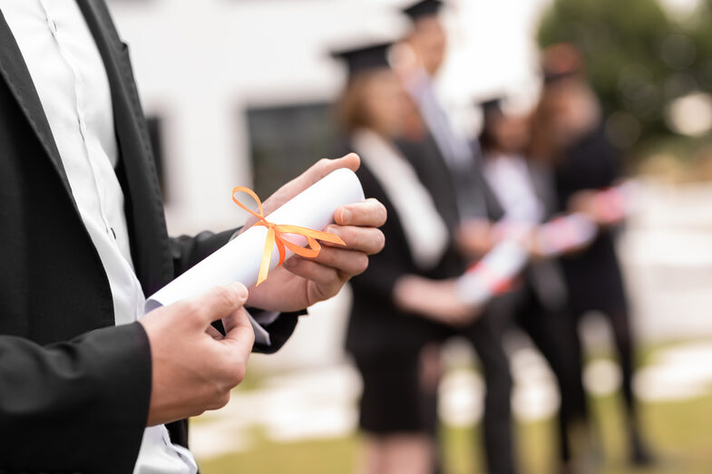 Photo of a rolled-up degree certificate with an orange ribbon held in someone's hand. In the background, people with doctor's hats can be seen very blurred.