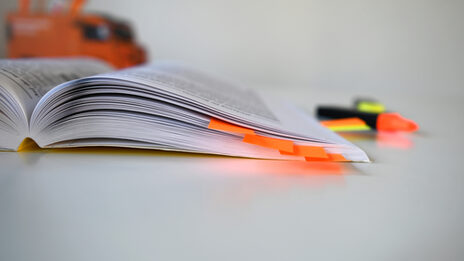 Foto von einem aufgeschlagenem Buch mit orangenen Notizzetteln drin. Im Hintergrund ist eine Stiftebox sowie Textmarker.