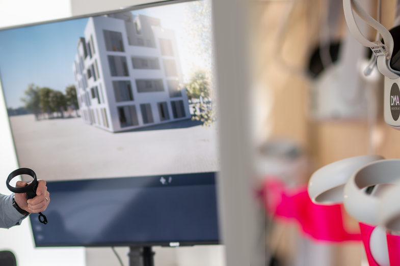 Foto von einem Mann mit VR-Brille auf und Controller in der Hand. Auf dem Bildschirm im Hintergrund sieht man ein Gebäude in der Perspektive, die auch der Mann durch die VR-Brille sieht. Rechts im Bild: Quest2 hängt an der 3D-gedruckten PEG-Board Halterung im VR Labor.