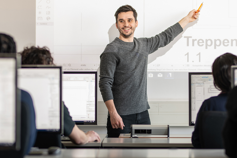 Foto eines Dozenten im Computerraum. Er zeigt etwas an einer Grafik, die auf die Wand projiziert wurde. Mit der anderen Hand zeigt er lächeln zu einem Studierenden, als würde er diese Person aufrufen. __ <br>An assistant professor is standing in the front of the computer room and is showing something on a graphic that has been projected onto the wall. With the other hand he shows a student smile as if he were calling that person.