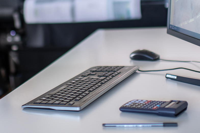 Zu sehen ist ein Bildanschnitt eines Arbeitsplatzes. <br>Rechts im Bild ist ein Teil eines Bildschirms, auf dem Schreibtisch liegen eine Tastatur, ein Taschenrechner, ein Stift und eine Maus.