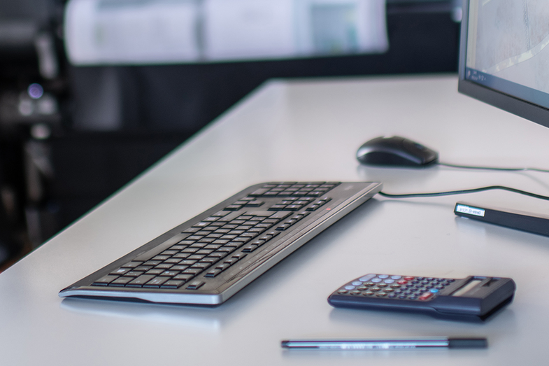 The picture shows a section of a workplace. <br>On the right of the picture is part of a screen, on the desk are a keyboard, a calculator, a pen and a mouse.