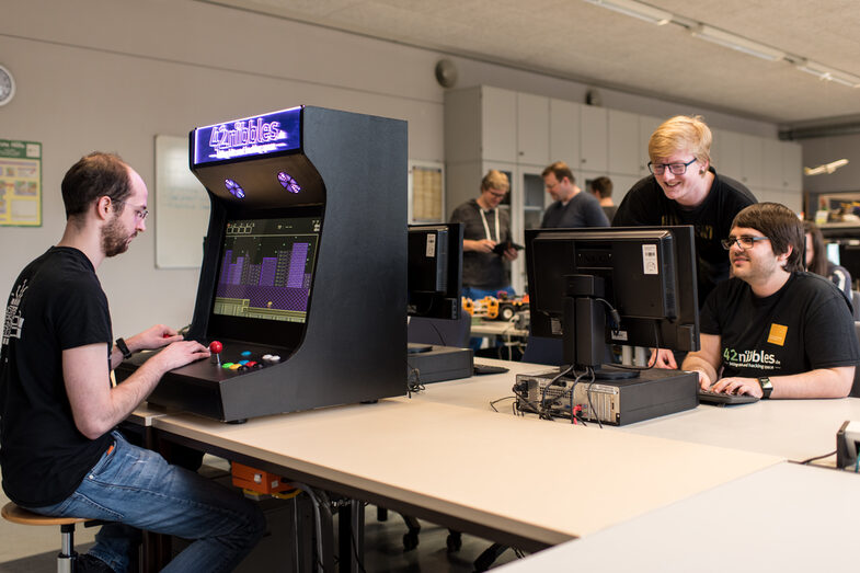 Foto vom offenen Labor des Fachbereichs Informatik. Ein Studierender sitzt links im Bild am selbstgebauten Spielautomat, in den hinteren Reihen des Labors sind zwei Studierende am Rechner im Gespräch sowie weitere Personen.