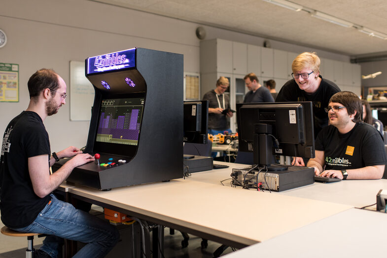 Photo of the open lab of the Faculty of Computer Science. A student is sitting at the self-built slot machine on the left of the picture, in the back rows of the lab two students are talking on the computer as well as other people.