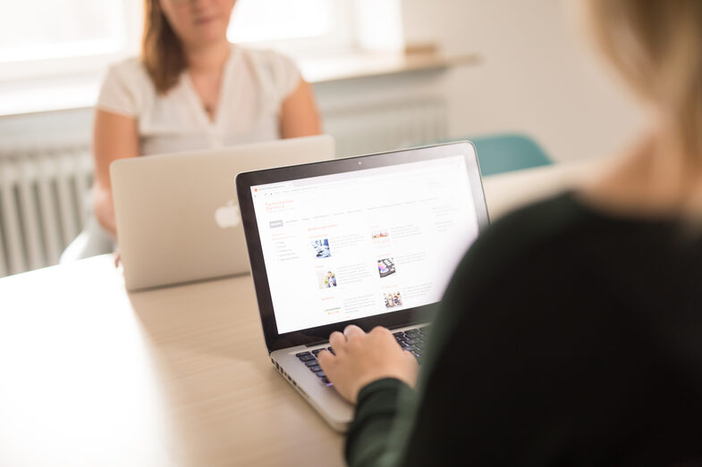 Photo of a young woman at the laptop, sitting across from her at the table, another woman with a laptop. __ Young woman at the laptop, sitting across from her at the table, another woman with a laptop.