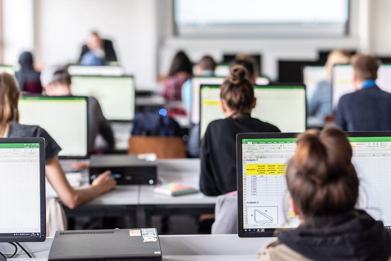 View from behind into the computer room with students, some Excel spreadsheets are visible on the monitors.