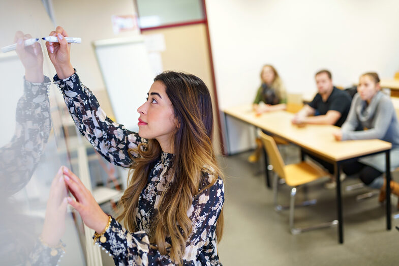 Seitliches Foto einer junge Frau, die etwas auf ein Whiteboard schreibt. Im Hintergrund unscharf zu erkennen Personen, die zuschauen.