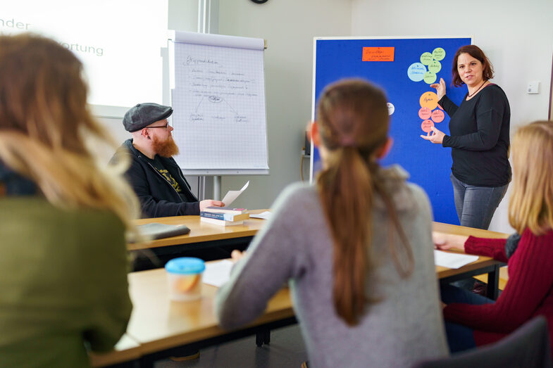 Photo of several participants of the ethnography working group sitting at tables. A woman stands at the front and explains something using a pinboard with several colorful pieces of paper.