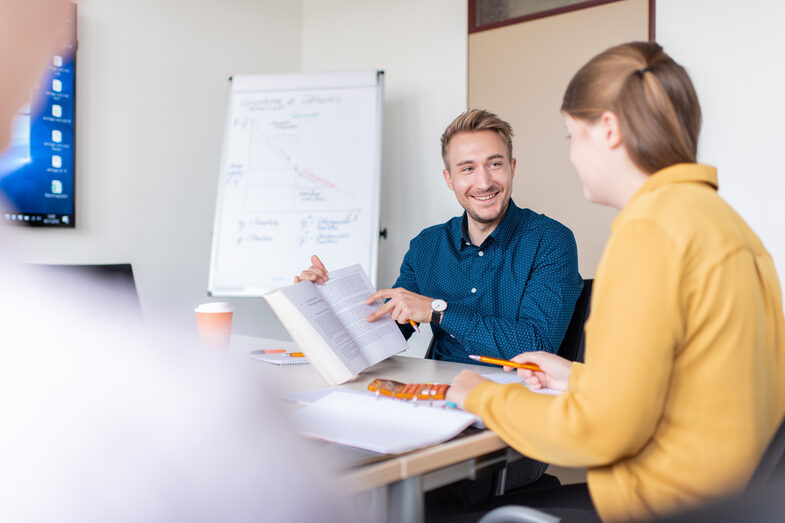 Three students are sitting at a table full of documents. One student is holding up a book and pointing at something while looking at the other student.