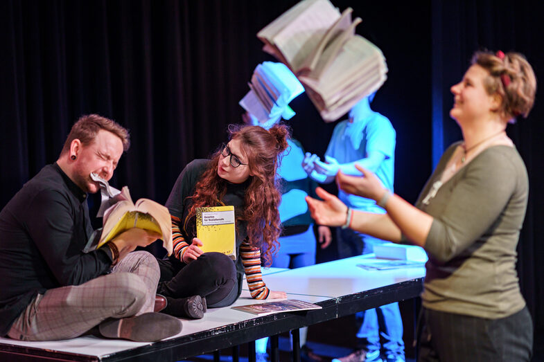 Foto von Schauspieler:innen, die eine Szene im Theaterlabor performen und dabei mit Büchern jonglieren.