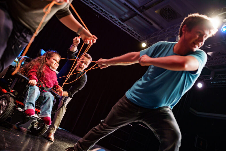 Photo from the Theaterlabor. Actors pull an actress in a wheelchair across the stage of the Theaterlabor using orange wool threads.