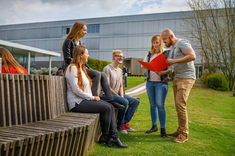 Foto von fünf Studierenden auf dem Campus. Zwei von ihnen schauen in eine Mappe, die anderen schauen ihnen zu.__Five students stand in front of and sit on benches on the campus. Two look into a folder, the other watch them.