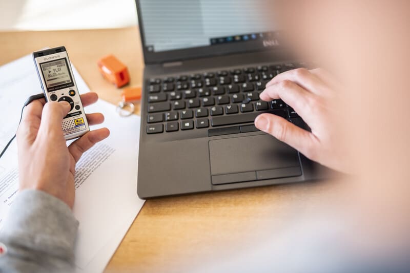 Foto eines Audio-Aufnahmegeräts, das von einer Person in der Hand gehalten wird, die Play-Taste wird betätigt. Daneben ein Laptop im Anschnitt, auf dem die Person tippt.