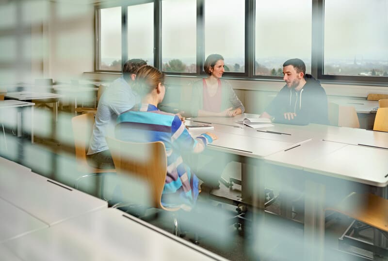 Foto durch eine Fensterscheibe in einen Seminarraum, in dem vier Studierende an einem Tisch sitzen und sich austauschen.