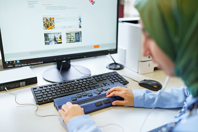 Foto der Hände einer Studentin mit Sehbehinderung, die auf der Braillezeile vor einem Computerbildschirm abgelegt sind.