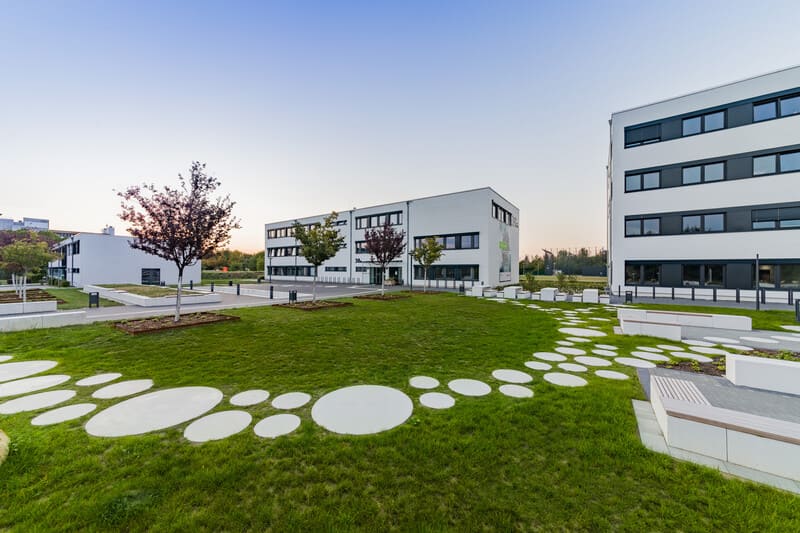 Foto des Gebäudes 38 a der Emil-Figge-Straße, daneben links das Gebäude 38 im Hintergrund und rechts im Anschnitt das Gebäude 38 b. Im Vordergrund ist die Wiese mit Bäumen sowie runde Bodenplatten als Gehweg.