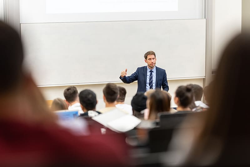 Lehrender steht im Hörsaal vorne vor seinen Studierenden und erläutert etwas.