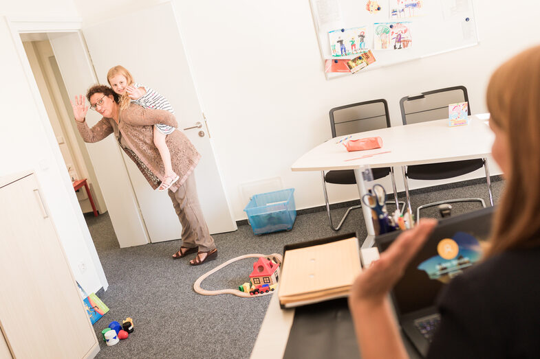 Photo of an office, with a woman sitting at a desk in the right-hand section. An older woman is standing in the doorway, carrying her little granddaughter piggyback. They wave to each other. There are children's toys on the floor.