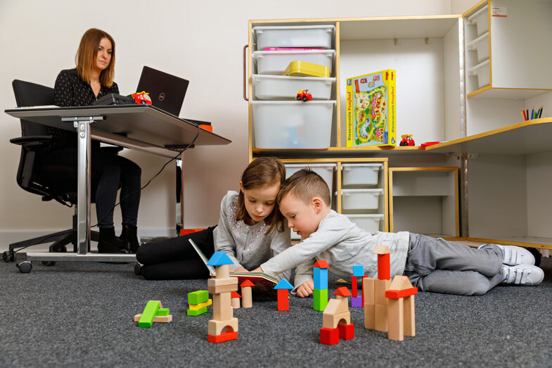 Foto einer Frau, die am Schreibtisch sitzt und arbeitet. Vor ihr spielen ihre Kinder auf dem Boden. Im Hintergrund steht die KidsBox. __ Woman sits at a desk and works - her children are playing on the floor in front of her. The KidsBox is in the background.