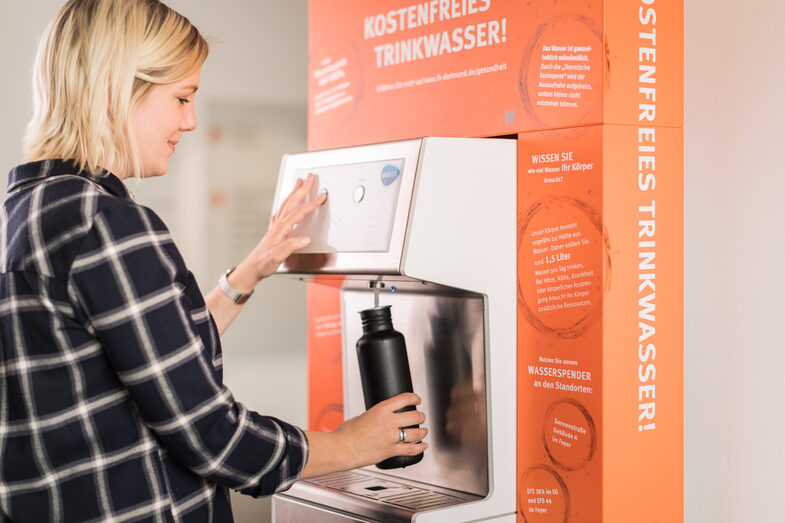 Foto von einer Studentin, die ihre Trinkflasche am FH-Wasserspender mit frischem Wasser befüllt.__Photo of a student filling her water bottle with fresh water at the water dispenser of the Fachhochschule Dortmund.