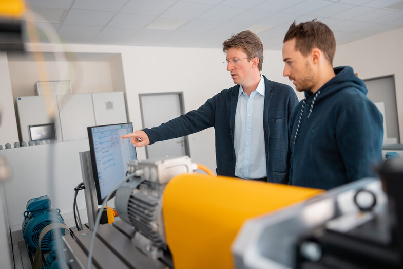 Foto von einem Professor und einem Studierenden, die hinter einem Elektromotorenprüfstand stehen. Der Professor erklärt etwas.