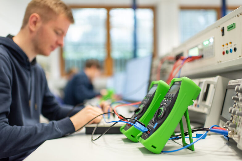 Photo of two measuring devices standing next to each other on the table and connected to another device with cables. Behind them, a trainee is working at his desk.