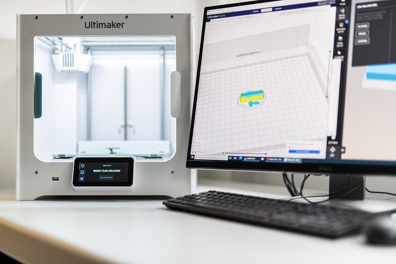 Foto eines Computerbildschirms, auf dem etwas für den 3D-Druck programmiert wird. Daneben ein 3D-Drucker mit quadratischer Form.