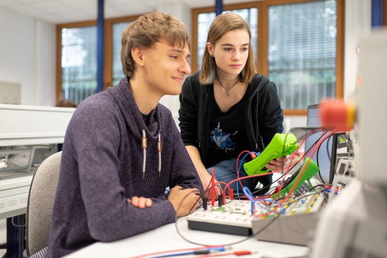 Foto von zwei Studierende im Labor für Elektronik und Automation. Sie sitzen an bzw. auf einem Tisch mit diversen elektronischen Messgeräten und schauen gemeinsam auf einen Bildschirm.