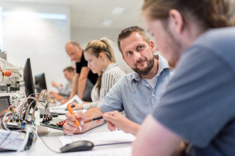 Foto von einem Professor, der mit einem Studenten im Labor für Elektronik und Automation im Gespräch ist. Sie sitzen an einem Arbeitsplatz mit elektronischen Versuchsaufbauten. Daneben weitere Studierende.