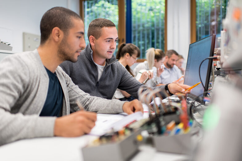 Foto von zwei Studenten im Labor für Elektronik und Automation an Arbeitsplätzen mit PC und elektronischen Aufbauten. Sie schauen gemeinsam auf den Bildschirm. Daneben weitere Studierende.